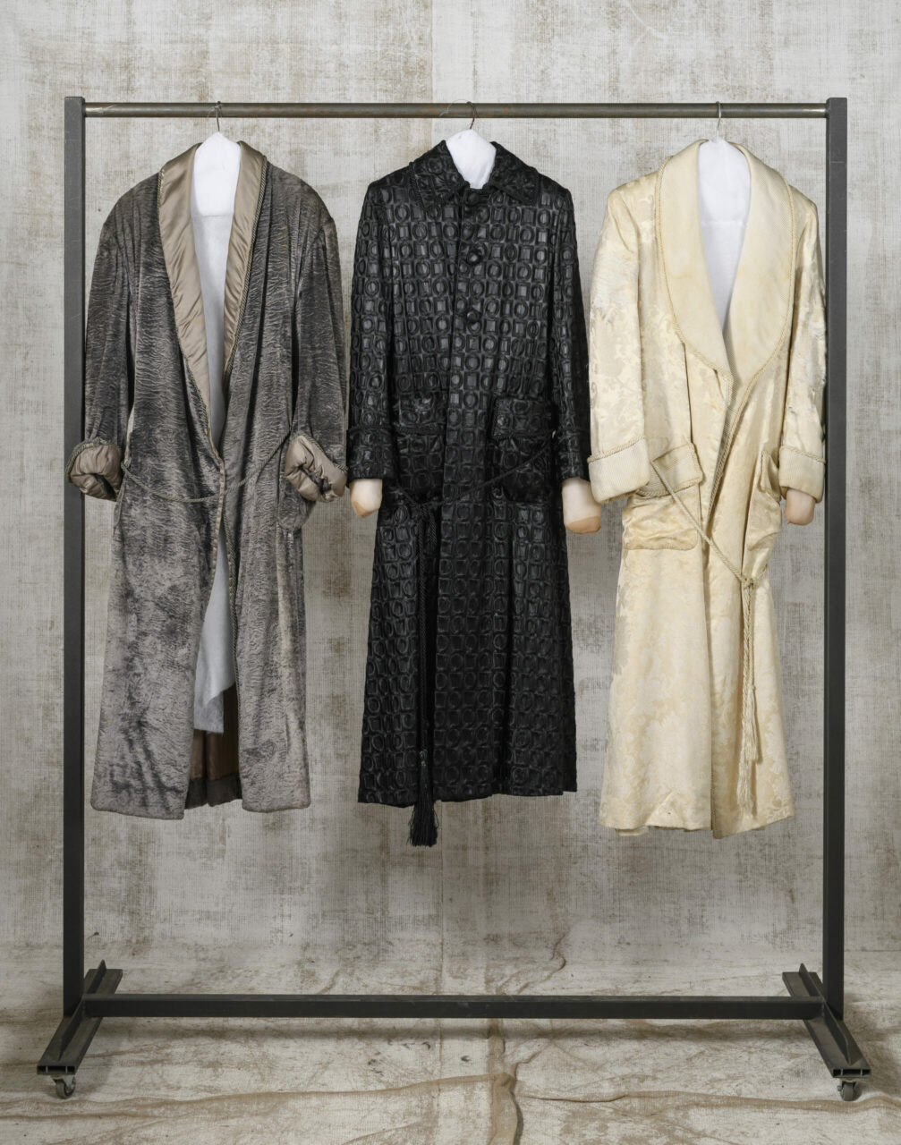 Three elegant bathrobes in different colors hang on a clothing rack against a neutral background wall.